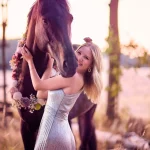 Junge Frau posiert im Wald neben Ihrem Pferd, dass mit eimem Kranz aus Blumen geschmückt ist